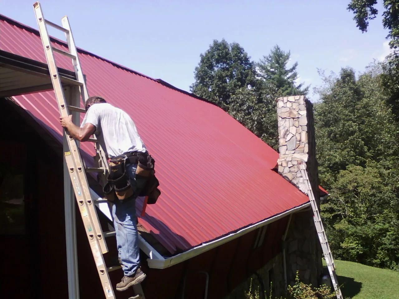 Roofer on a ladder beside a red metal roof and stone chimney — Soffit & Fascia Repair by Roofing Brothers in Kamas